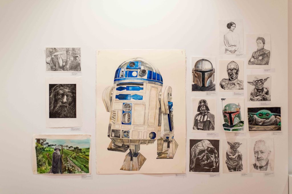 A collection of artistic works displayed on a wall, featuring a prominent illustration of R2-D2 in the center, surrounded by various portraits and sketches of characters from the Star Wars universe.
