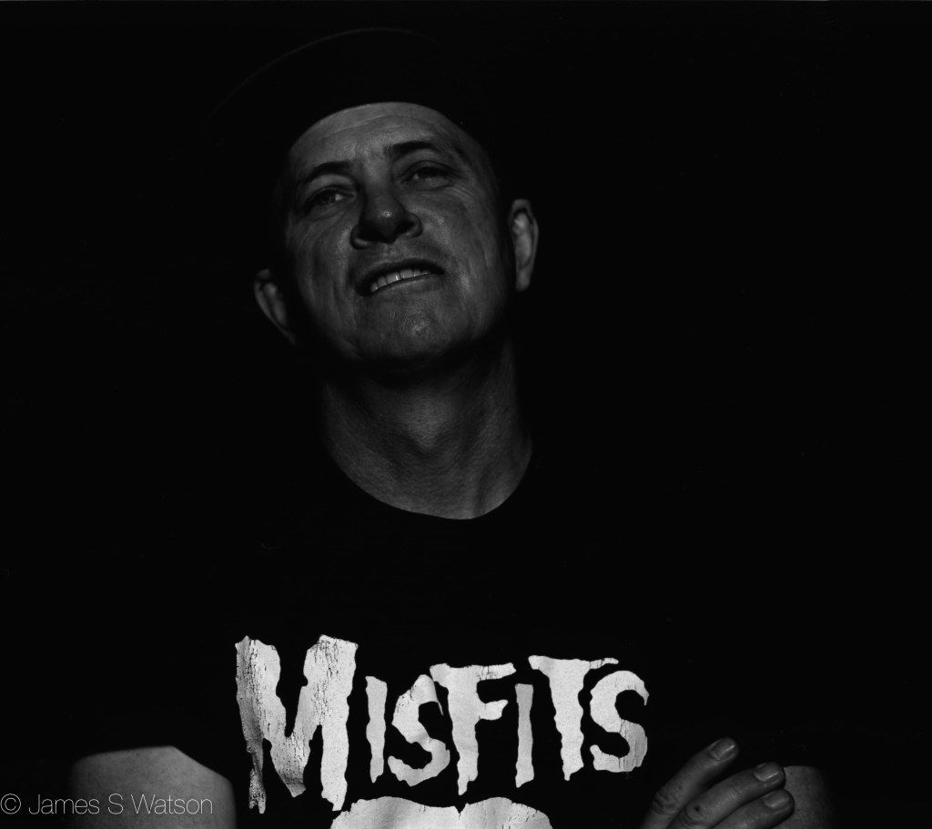 Black and white portrait of a man smiling, wearing a shirt with the word 'MISFITS' prominently displayed.